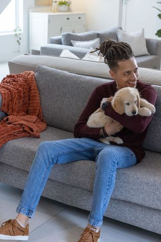 Smiling man hugging golden retriever puppy on modern grey sofa in sunlit living room