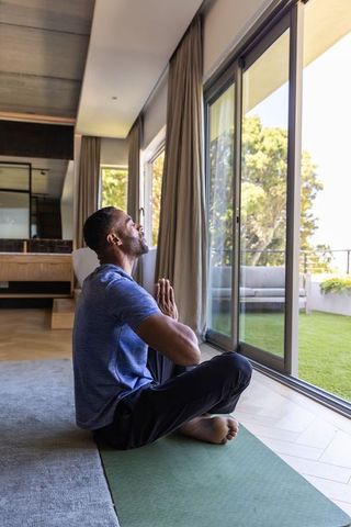 African american man meditating indoors on yoga mat near sunlit sliding glass doors