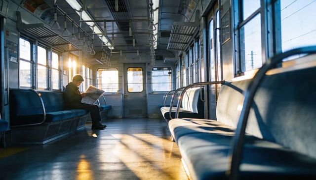 Reading commuter sitting alone on sunlit train carriage at sunrise, quiet urban commute