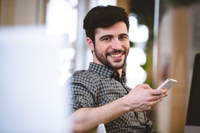 Young businessman smiling while using mobile phone in a modern, creative office environment. Ideal for use in business, technology, and communication-related content, showcasing a positive and relaxed work atmosphere.
