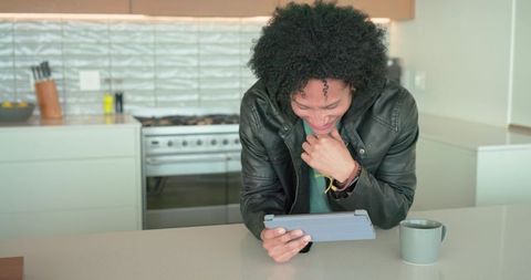 African american man leaning on kitchen island checking tablet and smiling over coffee