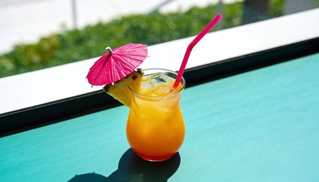 Sitting glass holding tropical orange cocktail with pink umbrella on turquoise table