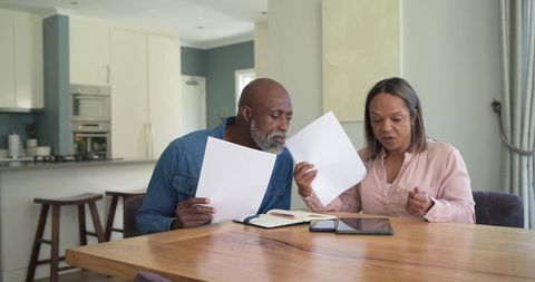 Mature couple reviewing financial documents at kitchen table with tablet and notebook