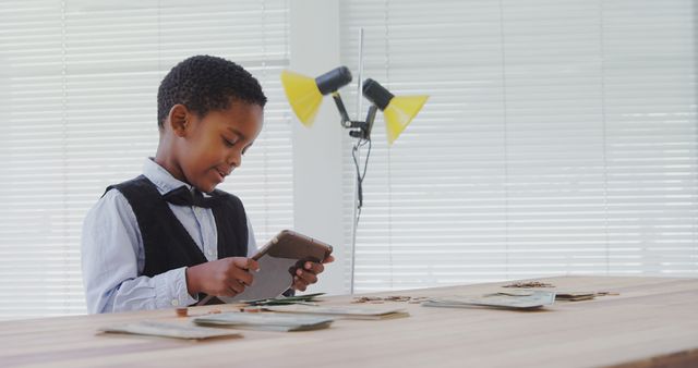 Young boy sitting at table counting money while using a tablet, bright natural light from windows. Ideal for themes related to childhood learning, early financial education, budgeting, and managing money at home.
