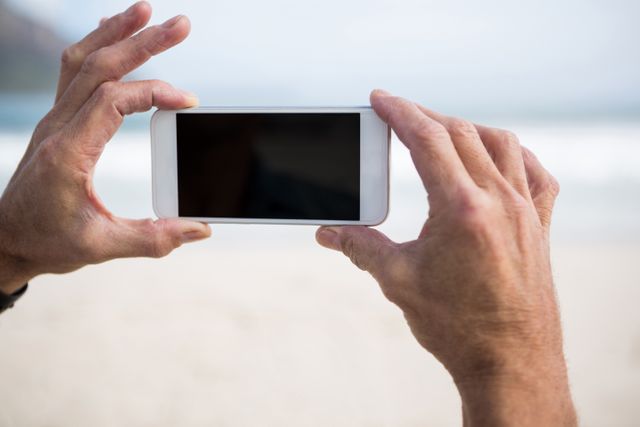 Close-up of hands holding a smartphone, capturing a picture on a beach during winter. Ideal for use in articles or advertisements related to travel, technology, photography, vacations, and outdoor activities.
