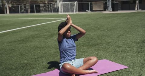 Black teen meditating on soccer field practicing outdoor yoga on purple mat