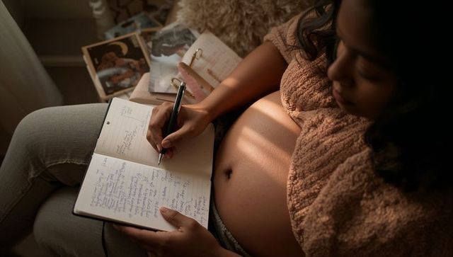 Pregnant woman journaling with planner on couch, sunlit bare bump and cozy knit sweater