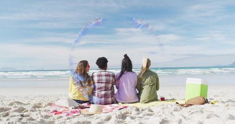 Four friends relaxing on sunny beach facing ocean with cooler, blankets and hats