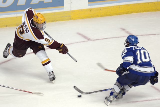 Two ice hockey players competing, each from different teams, on an ice rink. One player from the maroon jersey team is taking a shot, while the player from the blue jersey team is attempting to block. Suitable for use depicting athletic competition, team sports, ice hockey promotional materials, or articles on winter sports and athlete performance.