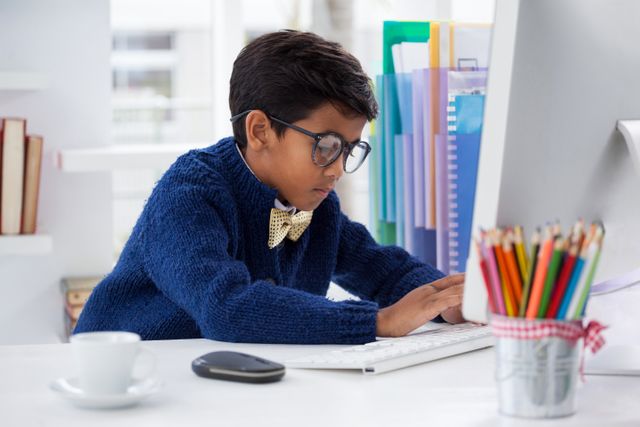 Young boy wearing glasses and a bow tie working on a desktop computer in a modern office. Ideal for use in educational materials, technology-related content, and articles about children in professional settings or learning environments.
