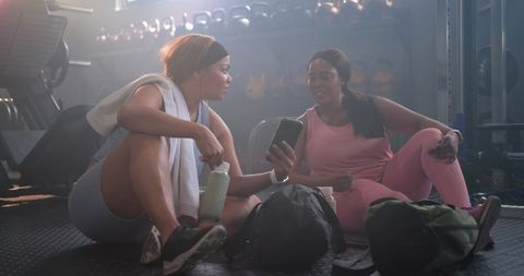 Diverse women relaxing and sharing phone after workout in gym