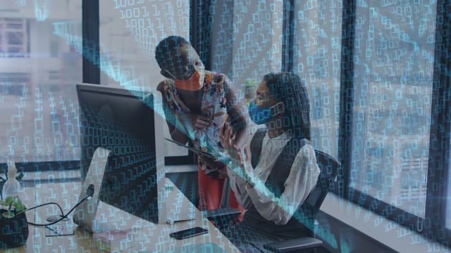 Two diverse female colleagues wearing face masks collaborate on a digital project in a modern office with overlaid binary code. This can be used for content related to teamwork in tech industries, pandemic workspace adaptations, and digital business solutions.