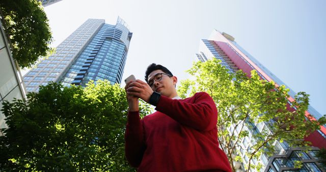 Young Asian man checks his smartwatch in an urban setting. Surrounded by skyscrapers, he stays connected while enjoying the outdoors.