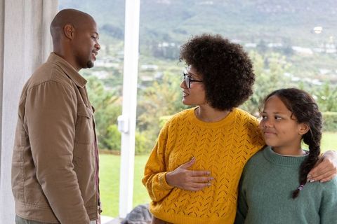 Diverse family standing by patio glass door, mother embracing daughter greeting father
