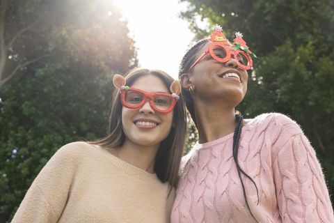 Diverse friends wearing festive novelty glasses standing in sunlit park smiling