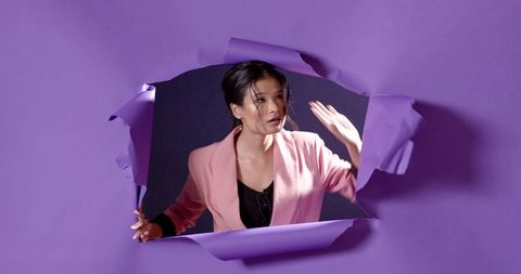 Asian woman emerging through torn purple paper wearing pink blazer dramatic portrait