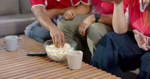 Diverse friends watching game on sofa, sharing popcorn and remote in casual sportswear