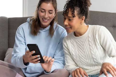 Diverse friends sharing smartphone and smiling while relaxing on sofa