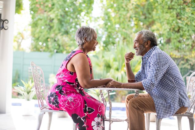 This image depicts a joyful senior couple sitting at a table in a garden, engaging in a lively conversation while holding hands. Ideal for use in advertisements, articles, or websites focused on senior living, retirement communities, healthy aging, relationships, and outdoor activities.