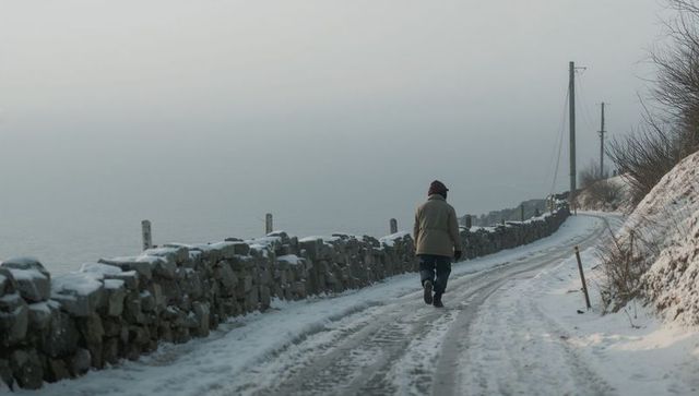 Senior walking alone along snowy coastal lane with dry-stone wall and misty horizon
