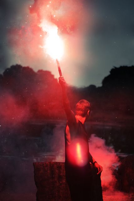 Dramatic moment with a person holding a red flare, emitting smoke against a nighttime backdrop. Suitable for themes of adventure, exploration, mystery, and night-time activities. Can be used in marketing for outdoor gear, adventure travel, or artistic projects requiring a dramatic and intriguing atmosphere.
