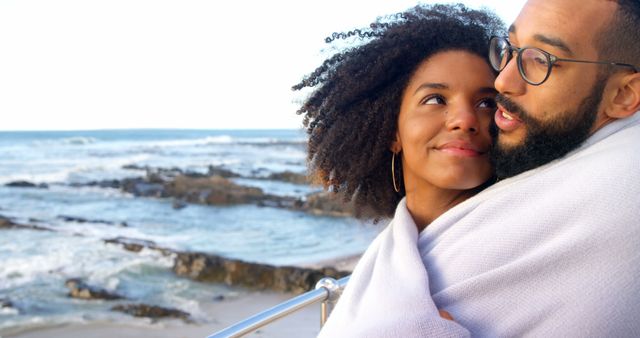 Couple lovingly embracing by the ocean, wrapped in a light blanket. Useful for themes of love, romance, togetherness, outdoor activities, vacations, and romantic getaways.