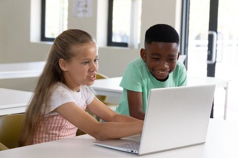 Multicultural children collaborating on laptop in bright classroom learning digital skills