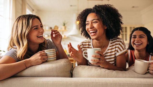 Laughing and chatting friends holding coffee mugs on couch in bright cozy kitchen