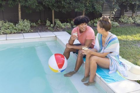 Diverse couple relaxing by poolside dipping feet and sipping drinks on summer afternoon