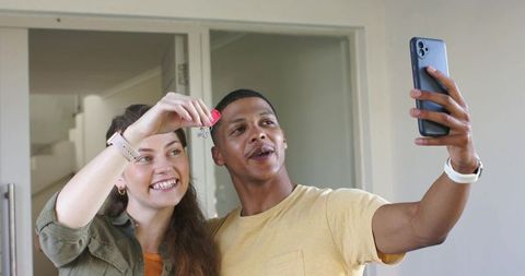 Young couple celebrating new home holding keys and taking joyful selfie in entryway