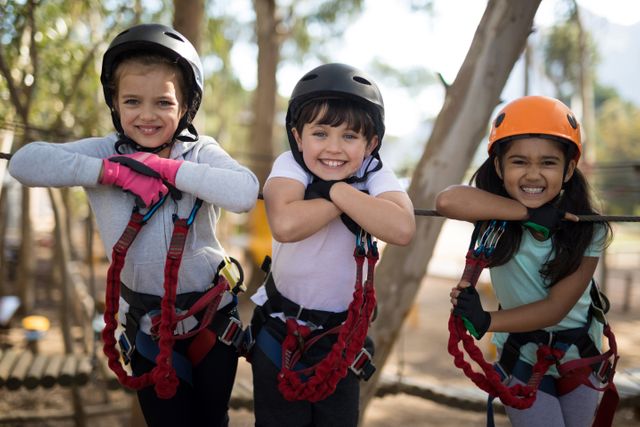 Three children are smiling and leaning on a cable at an outdoor adventure park. They are wearing helmets and safety harnesses, indicating they are participating in a fun and safe activity. This image is perfect for promoting outdoor activities, summer camps, team-building exercises, and childhood adventure experiences.