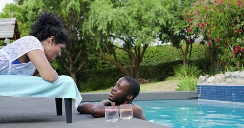 Diverse couple chatting at backyard pool, woman leaning on lounge, relaxed summer moment