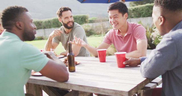 Happy diverse male friends sitting at table laughing, eating and drinking. Spending time together outdoors.