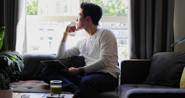 Pensive Young Man Sitting on Couch Looking Out Window Holding Tablet - Download Free Stock Images Pikwizard.com