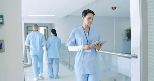 Image shows a healthcare professional using a tablet in a hospital corridor. The nurse is wearing medical scrubs and is focused on the tablet, possibly checking patient information. Suitable for articles or content focused on technology in healthcare, nursing profession, or hospital environments.