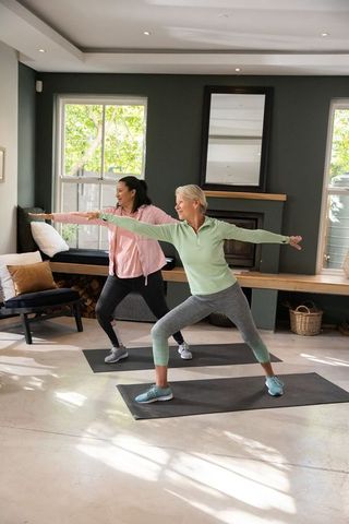 Senior women practicing warrior ii yoga on mats in sunlit cozy living room with fireplace