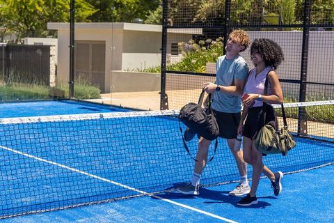 Couple walking across vibrant blue padel court carrying gym bags, water bottle, phone