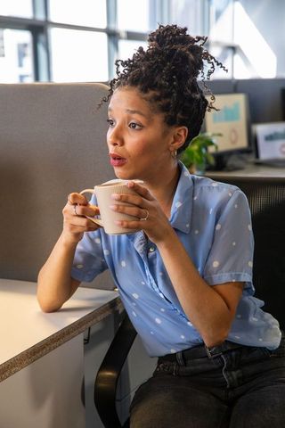 African american woman holding mug, glancing at monitors during break in open-plan office