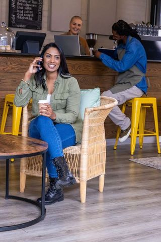 Smiling woman talking on phone while holding coffee in cozy multicultural café interior