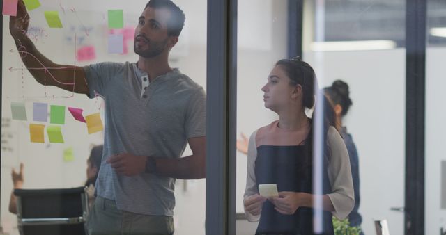 Two colleagues, a man and a woman, brainstorm with sticky notes on a glass board in a modern office. This image is ideal for illustrating team collaboration, innovative planning processes, business strategies, and dynamic work environments in articles, blogs, corporate websites, marketing materials, and startup promotions.