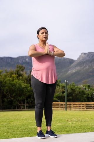 Asian woman practicing standing prayer pose in mountain park with pink top and smartwatch