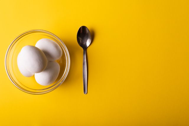 Overhead view of white eggs in a glass bowl next to a spoon on a vibrant yellow background. Ideal for use in food blogs, cooking websites, healthy eating promotions, and kitchen-related advertisements. The bright yellow background adds a cheerful and modern touch, making it suitable for various culinary and health-focused content.