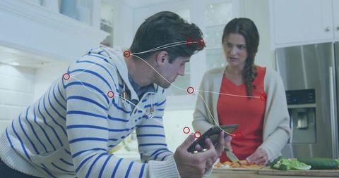 Couple meal prep with man checking phone and woman chopping vegetables in modern kitchen