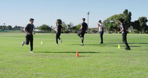 Diverse male athletes running cone-drill warmup on sunlit grass field with coach guiding