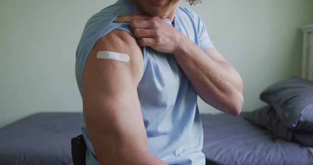 Man wearing light blue t-shirt showing bandage on upper left arm after receiving vaccination. Bedroom background with bed visible. Useful for healthcare promotions, vaccination campaigns, health awareness materials.