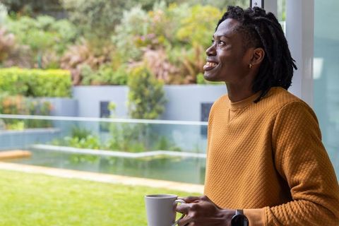 Young man holding mug at glass door overlooking backyard pond, smiling and contemplating