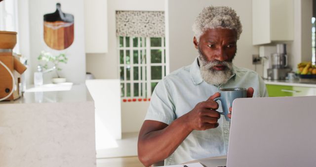 A mature man with gray hair and a beard is drinking coffee while using a laptop in a modern kitchen. This can be used for topics related to remote work, casual lifestyle, daily routines, home office setup, and technology use by seniors. Ideal for blogs, articles, and advertisements focusing on working from home, healthy routines, or lifestyle for older adults.