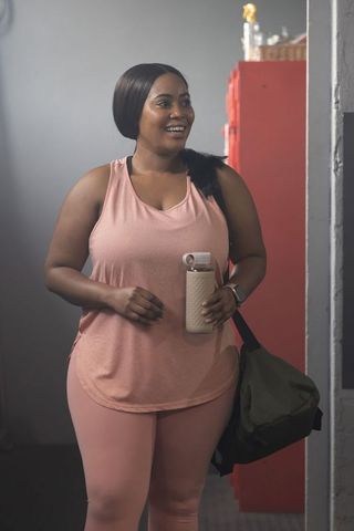 African american woman in pink smiling in locker room holding water bottle and duffel
