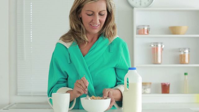 A woman is pouring milk into a bowl of cereal in her kitchen while wearing a green robe. The kitchen has a bright atmosphere with shelves holding various containers in the background. This video can be used for promoting healthy eating, morning routines, and lifestyle content.