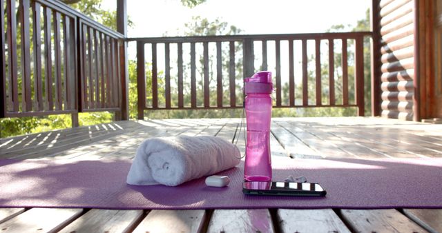 This serene scene depicts an outdoor yoga mat with a rolled-up towel and a pink water bottle on a wooden deck. A smartphone and white earbuds are placed next to the water bottle, giving the impression of a relaxing yoga session or workout in nature. The background shows greenery and the peaceful environment, making it ideal for promoting wellness, fitness routines, personal health blogs, and nature-related activities.